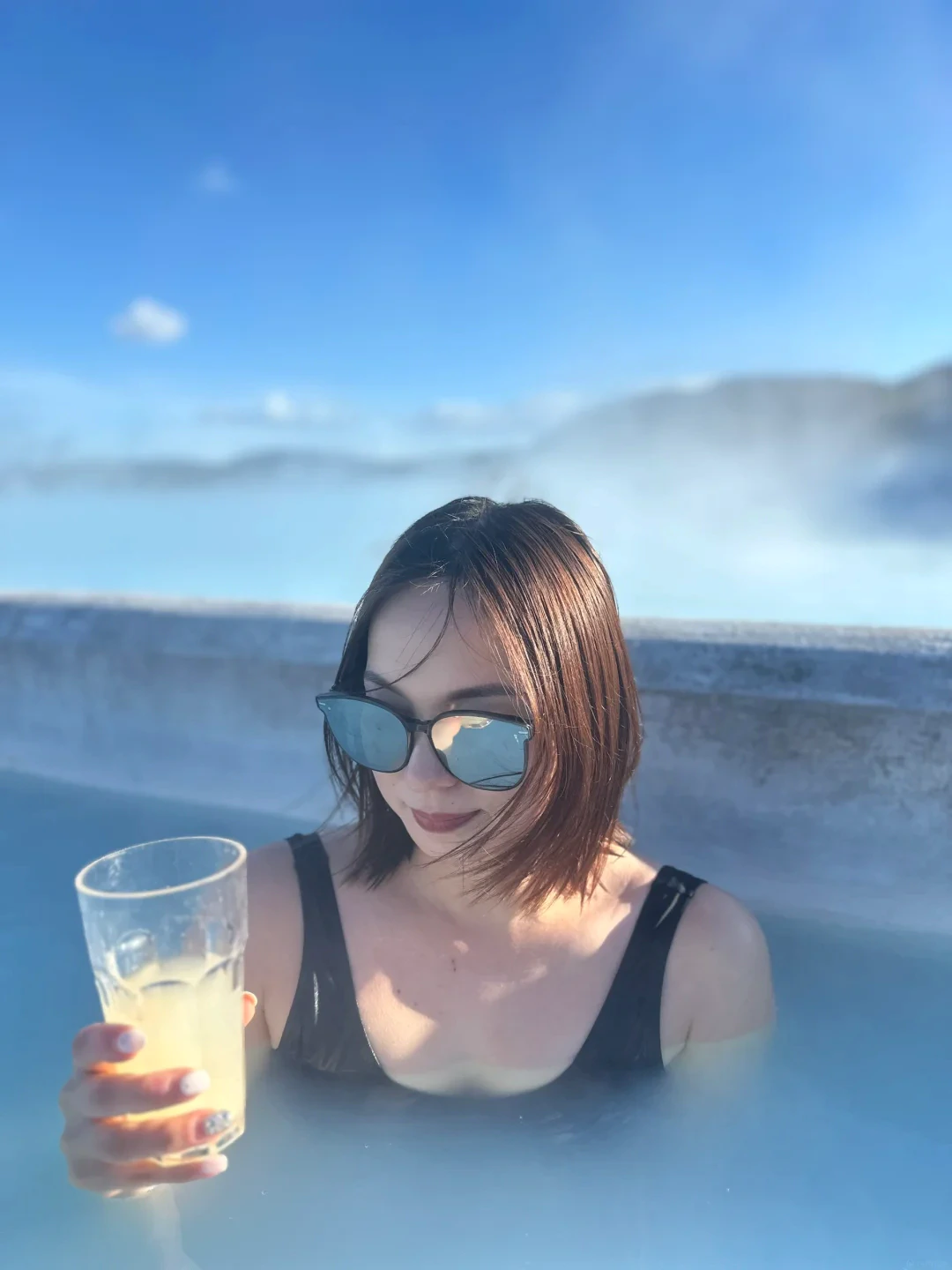 Alicia in sunglasses holding a drink in a blue Icelandic geothermal lagoon